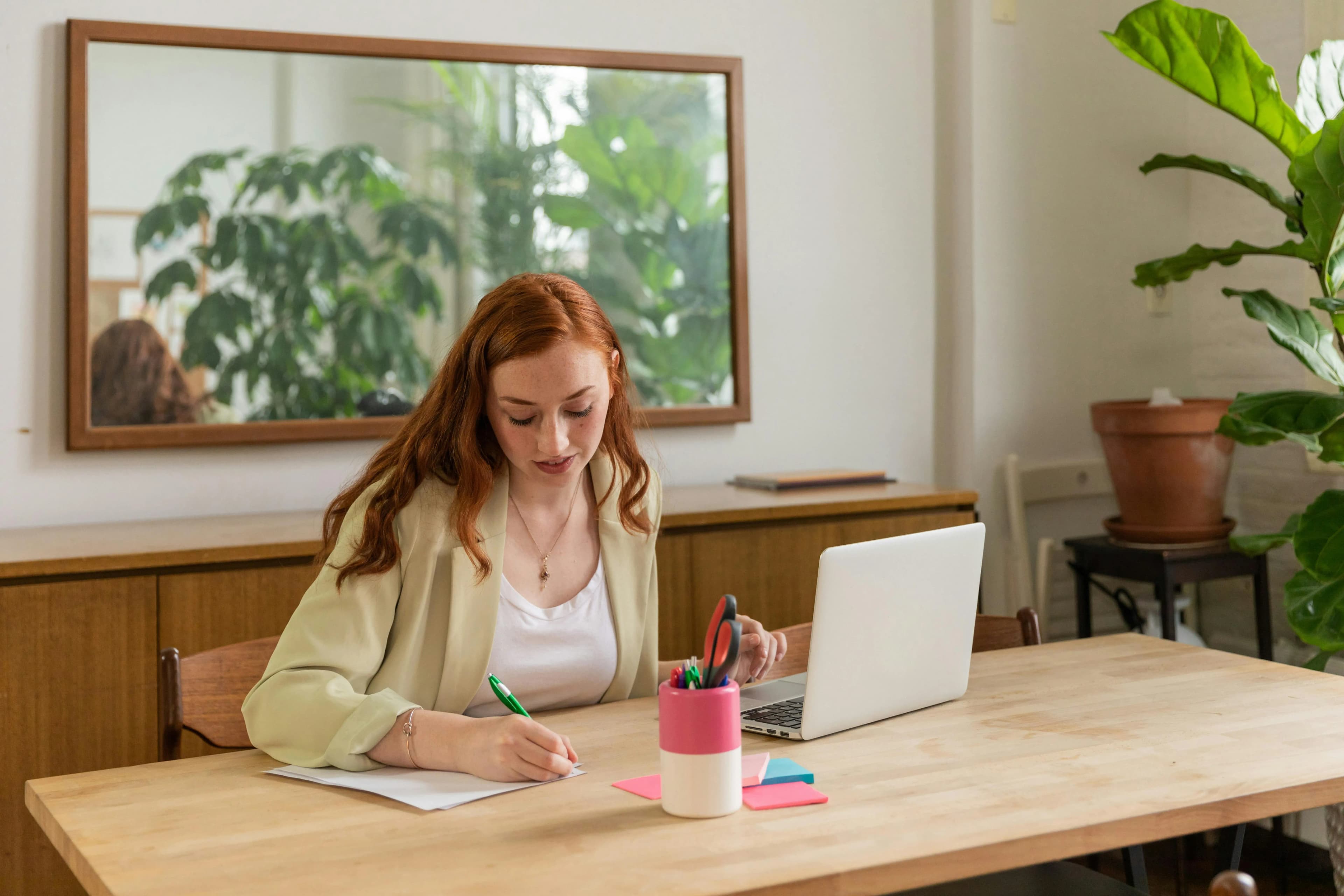 Woman sitting at a table writing on a piece of paper,  she also has her laptop open.