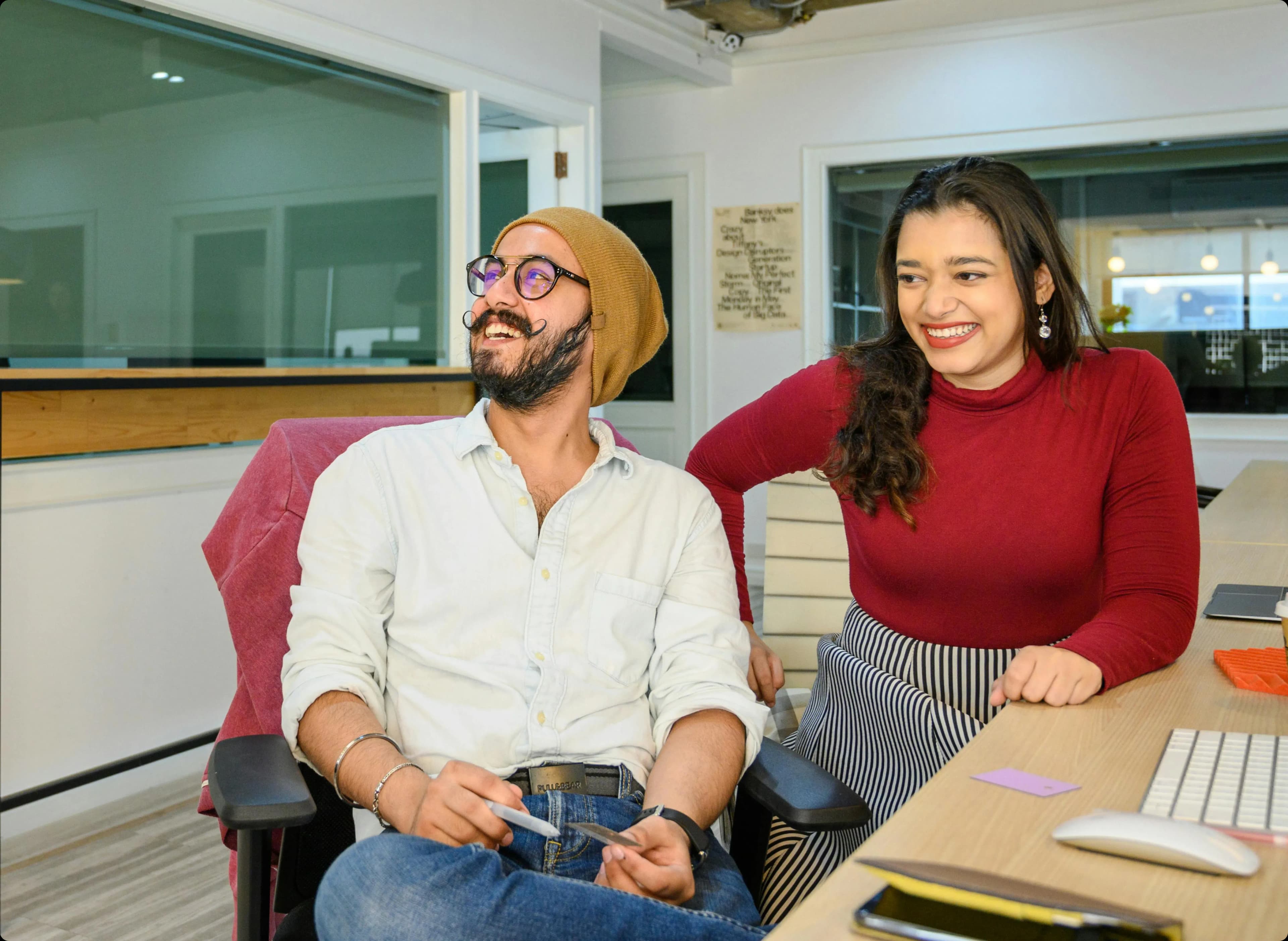 Two office workers sitting at desk facing away from each other, both smiling