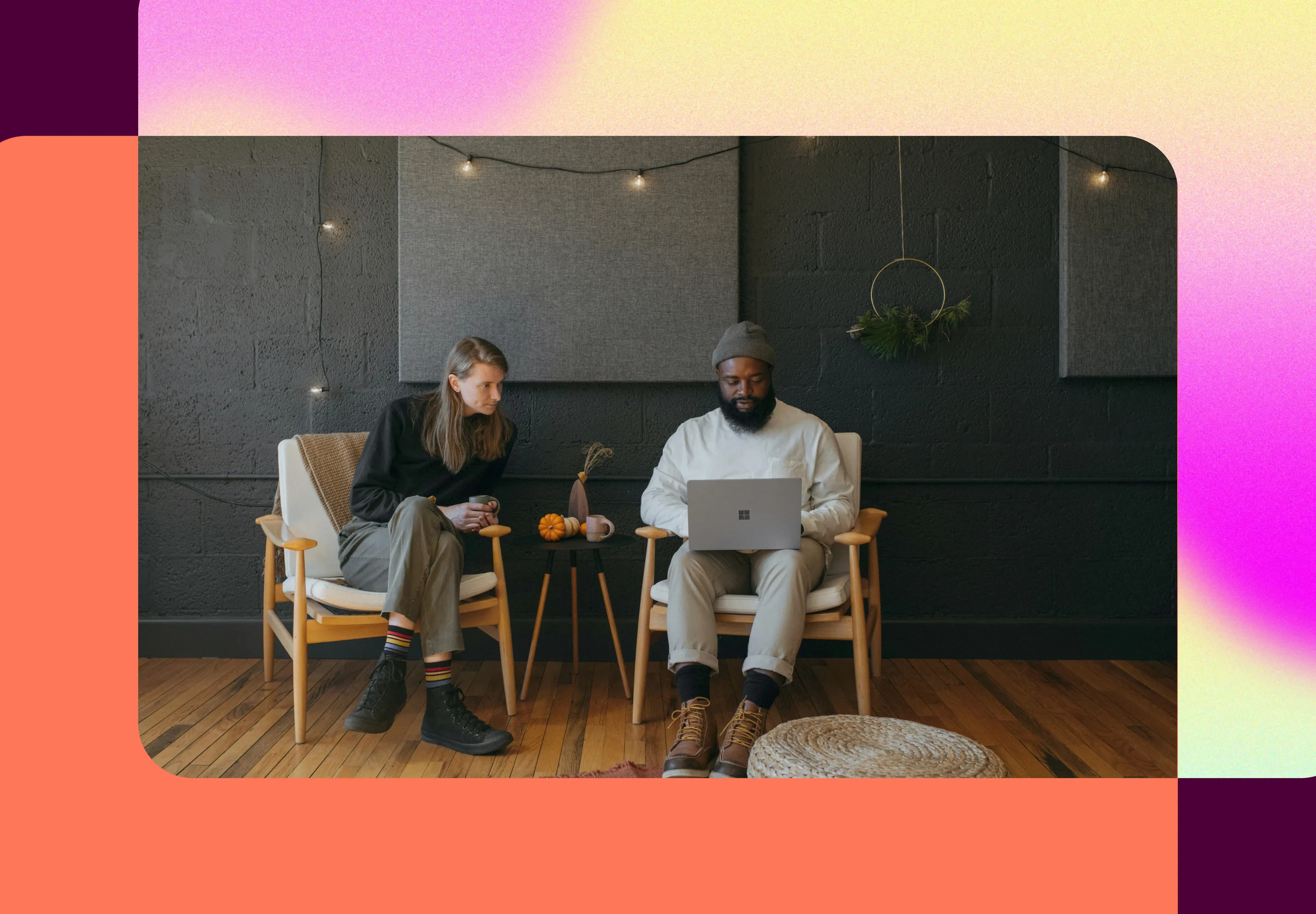 Two people sitting on chairs in a cozy room with string lights, using a laptop on a small table, with a colorful gradient border.